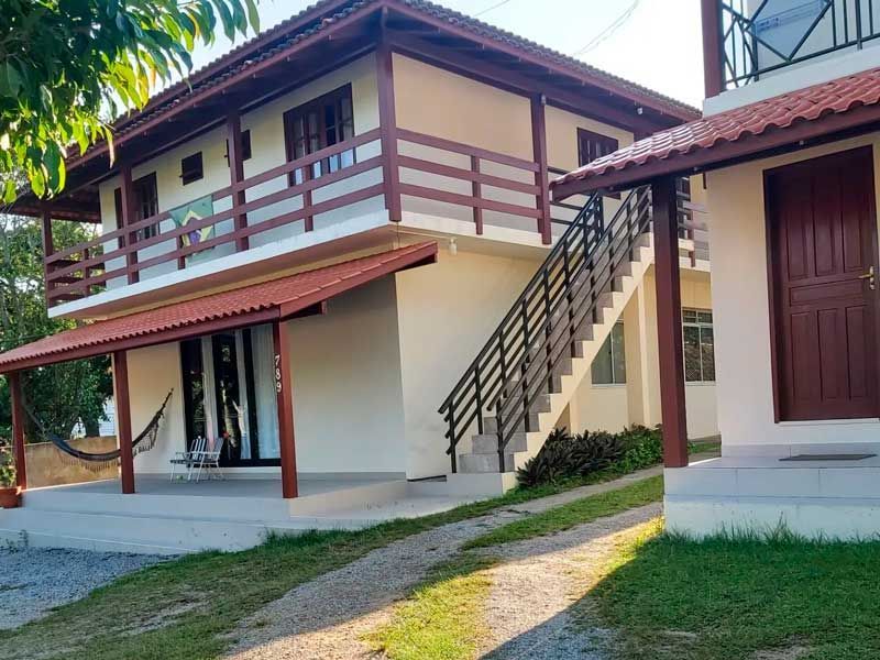 Prédio de dois andares, cor creme, com detalhes em marrom, escadas e telhado vermelho, localizado na área externa.