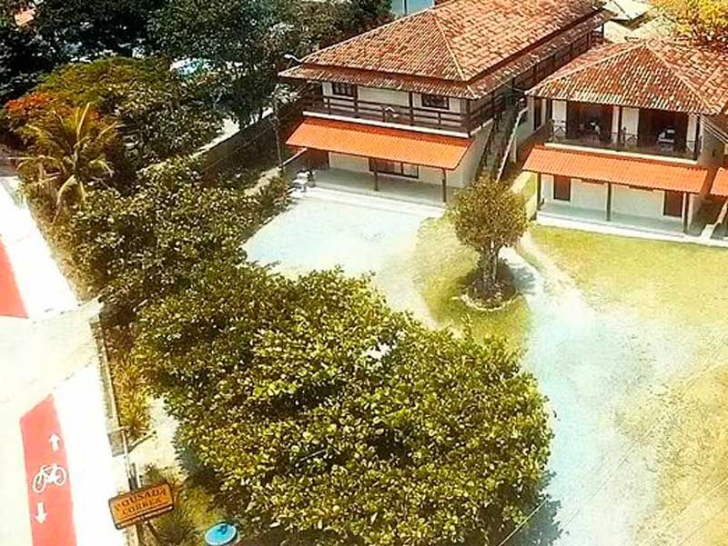 Vista aérea de um edifício de dois andares com telhado de telhas vermelhas, rodeado por vegetação e um pátio pavimentado.