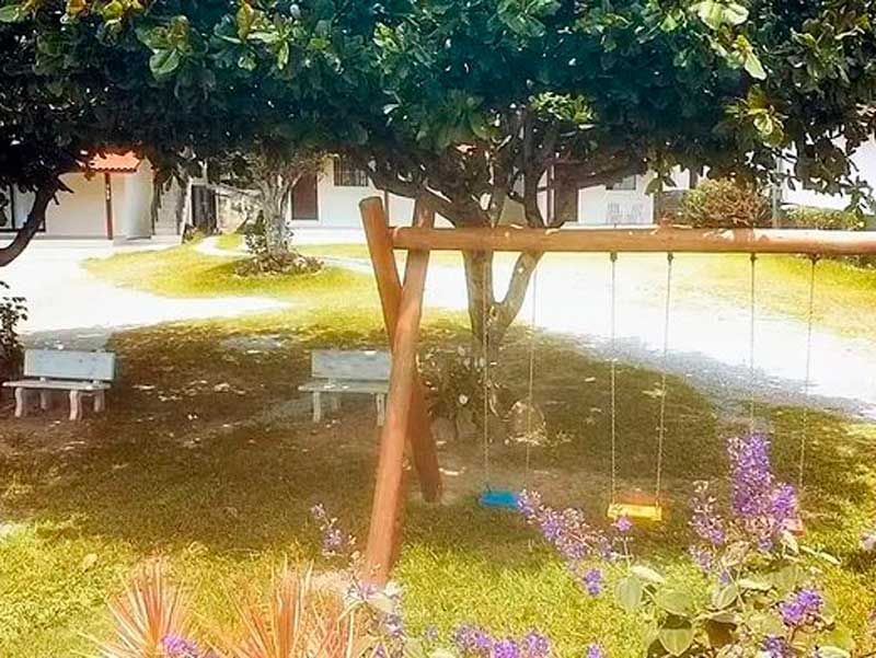 Um balanço de madeira em frente a uma árvore e casas; flores roxas e bancos em uma área gramada.