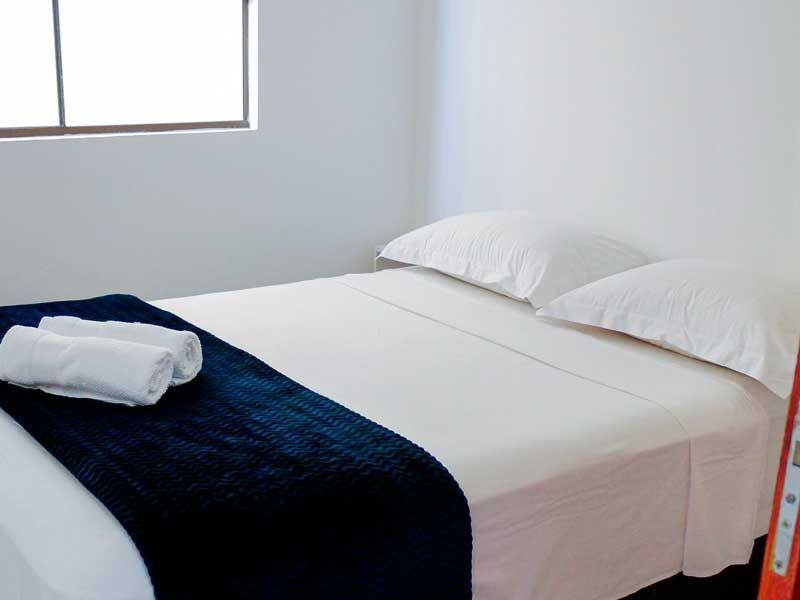 Quarto com roupa de cama branca, cobertor azul-marinho, toalhas enroladas e uma janela.