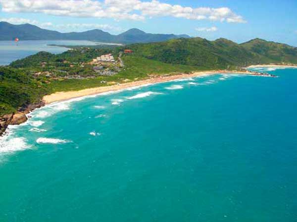 Águas costeiras azul-turquesa encontram uma praia de areia branca, emoldurada por colinas verdejantes sob um céu azul.