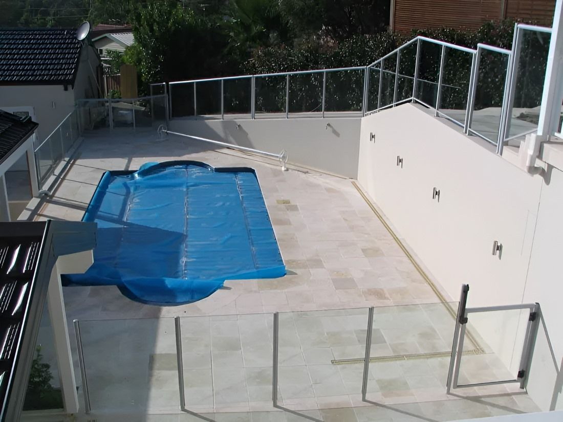 Outdoor pool with blue cover, surrounded by light stone patio, white walls, and glass railings — D & T Balustrade Systems In Unanderra, NSW
