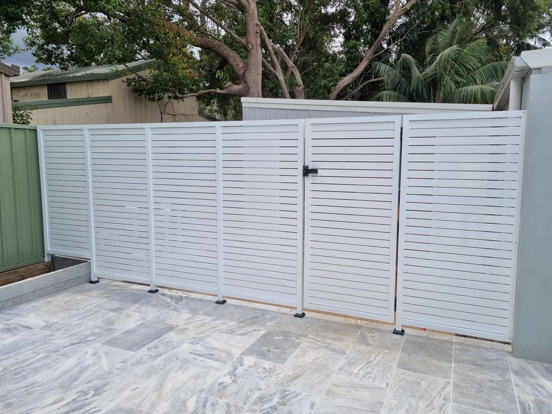 White slatted metal fence and gate on a paved area, trees and building in the background — D & T Balustrade Systems In Unanderra, NSW