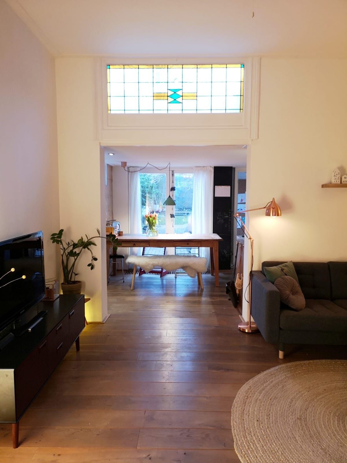 Woonkamer met zicht op de eetkamer; glas-in-lood boven de deur. Houten vloer, donkere bank, rond tapijt.