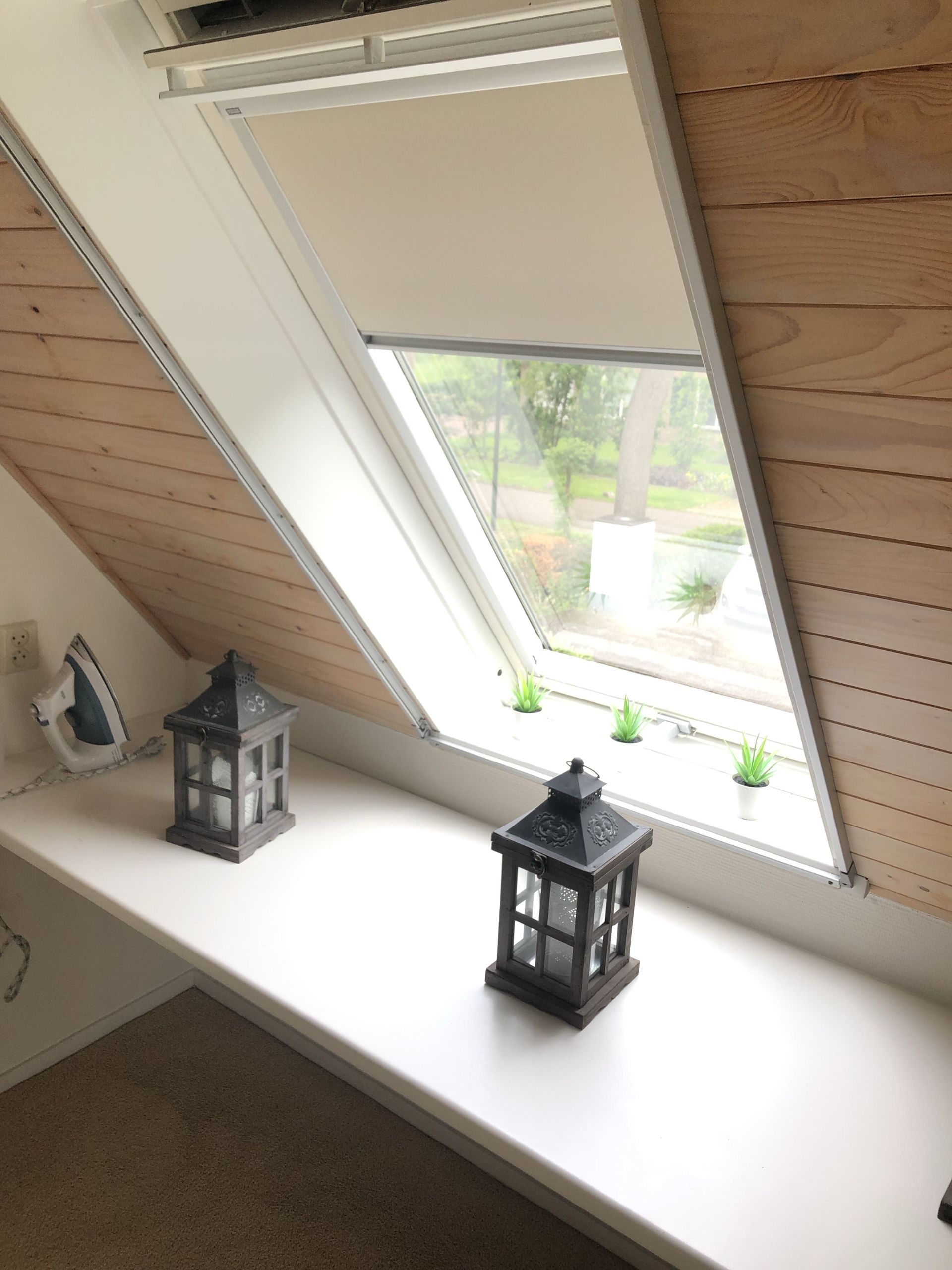 Schuin raam met gesloten gordijn, lantaarns en een strijkijzer op een witte vensterbank met houten lambrisering.