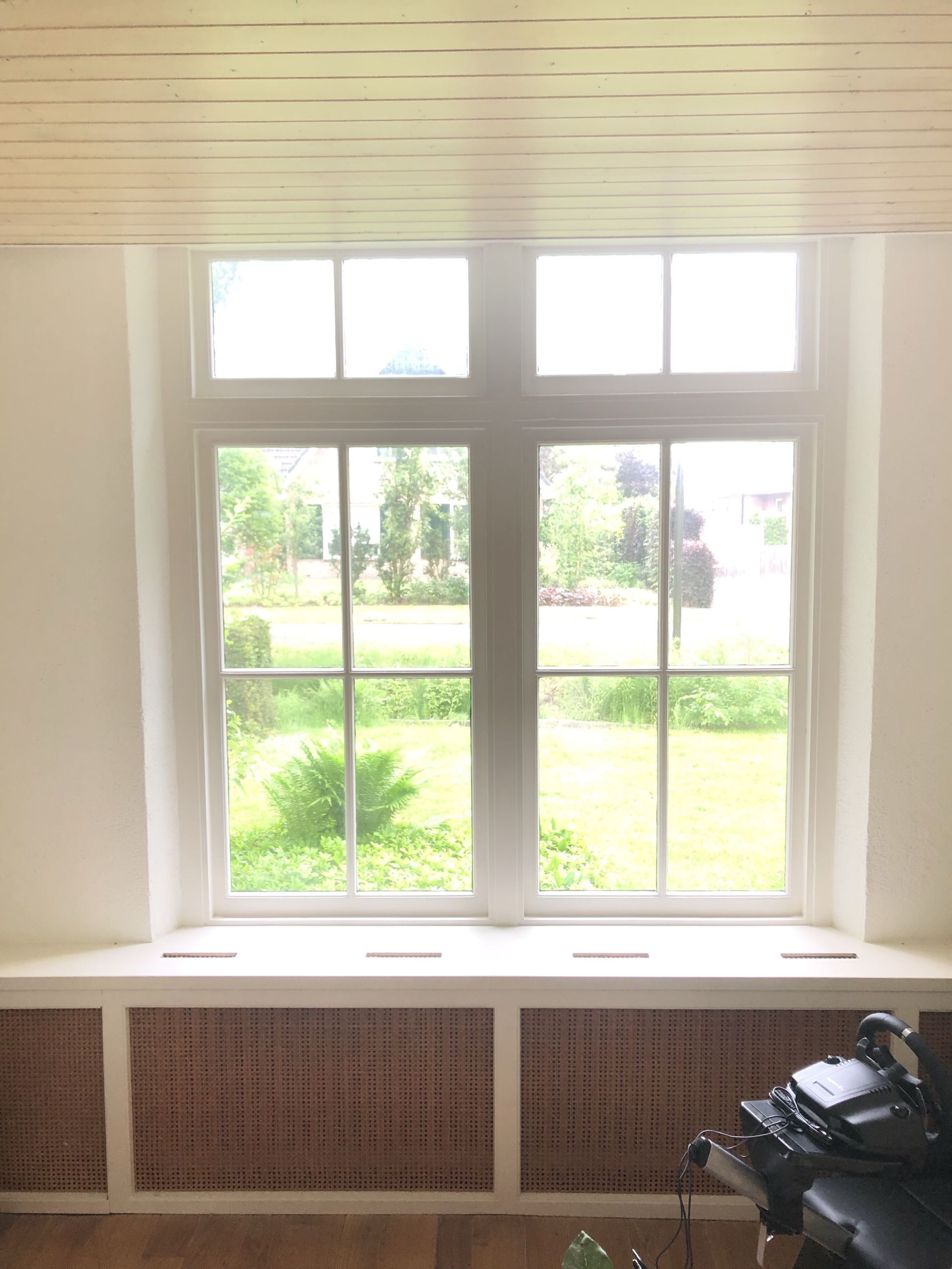 Wit raam met meerdere ruiten, uitkijkend op een groene tuin. Daaronder een bruin traliewerkpaneel.