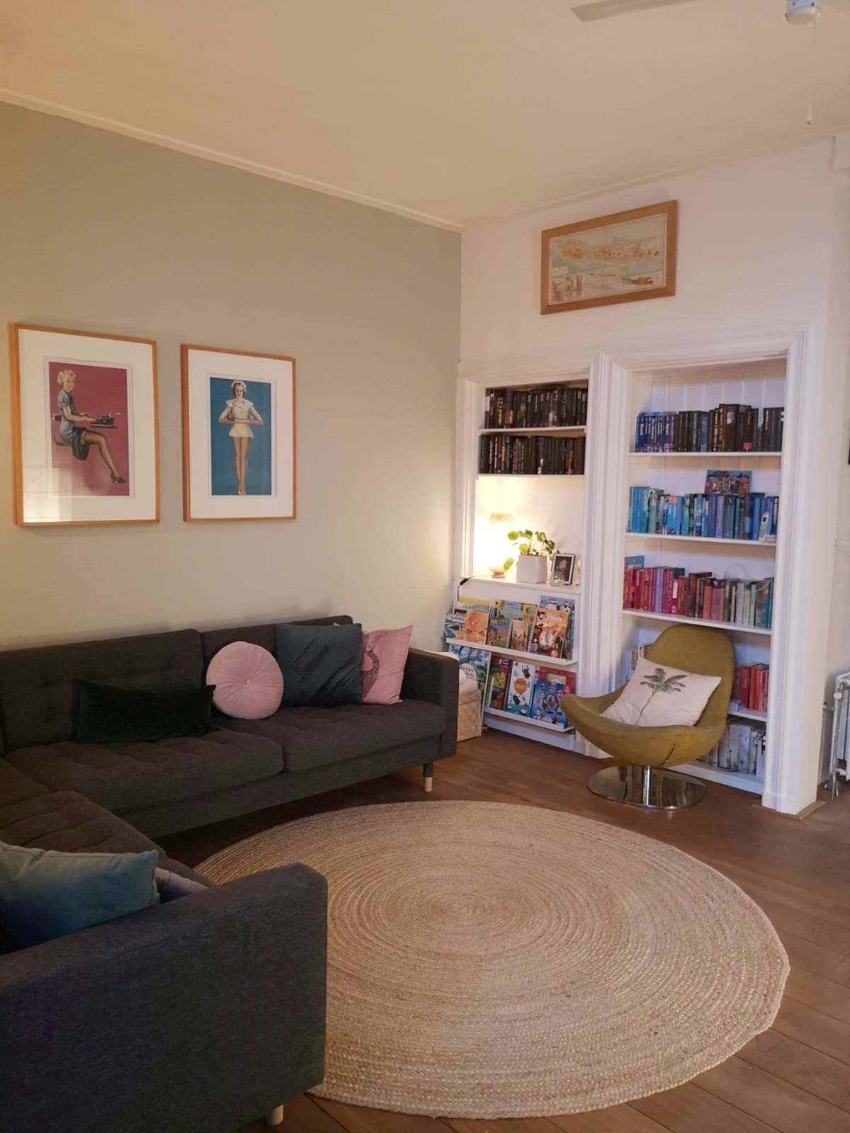 Woonkamer met grijze bank, rond vloerkleed, ingebouwde boekenplanken en ingelijste kunstwerken.