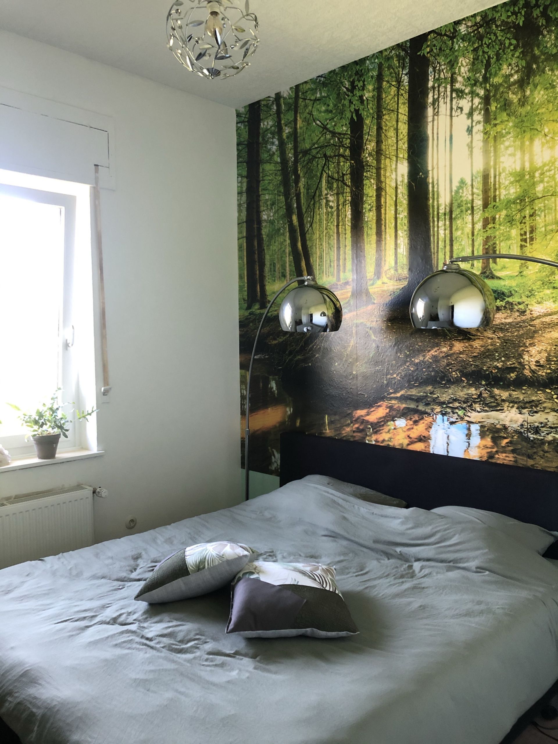 Slaapkamer met hoofdeinde met muurschildering in bosmotief, bed met grijs linnen, raam met witte rand en chromen lampen.