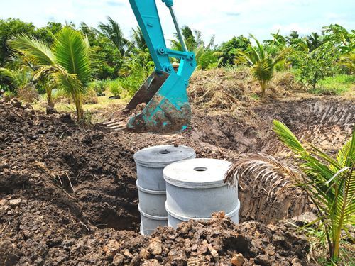 An excavator digs soil near two concrete septic tanks in a tropical area with palm trees.
