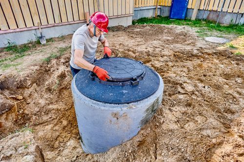 A plumber installs a concrete septic tank lid in an outdoor dug-out area with dirt and grass.