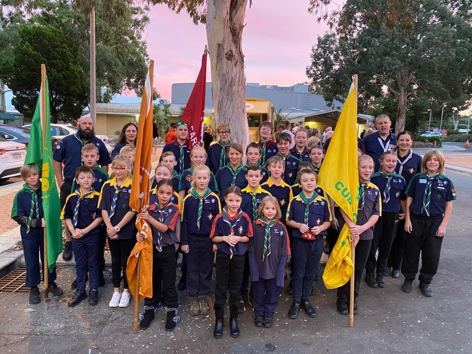 Scouts in uniform holding flags, standing in a group outdoors. Dawn sky, mixed ages, smiling faces, ANZAC Service.