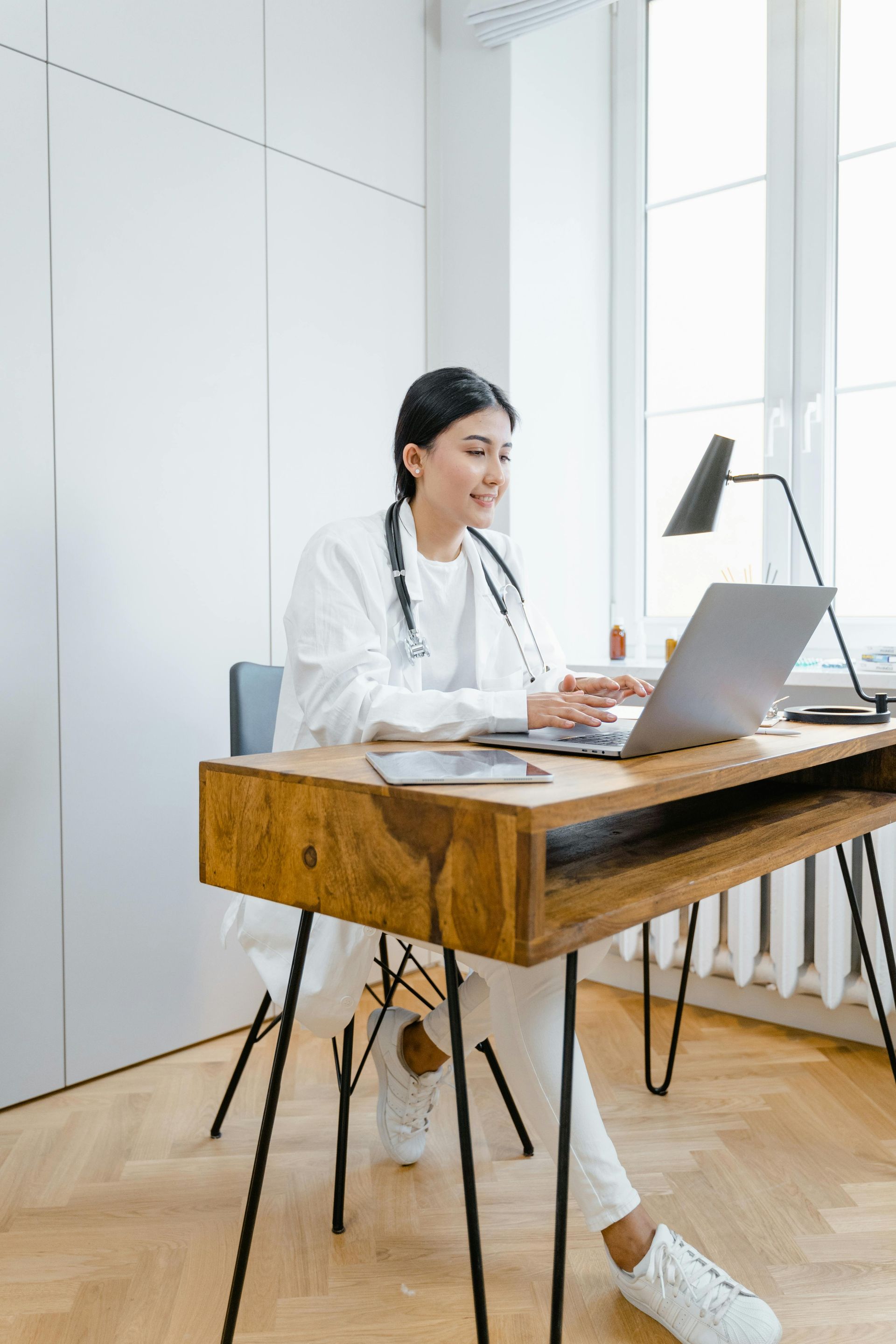 A female doctor is sitting at a desk using a laptop computer.