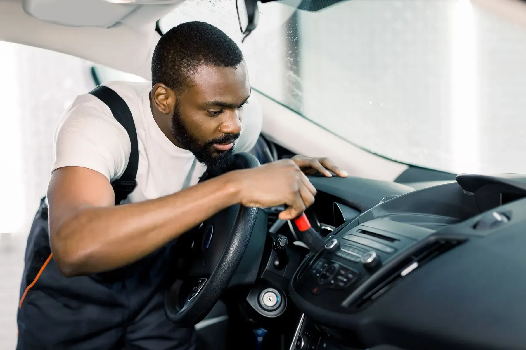Mechanic cleaning a car dashboard with a brush. Black man in work overalls inside a vehicle.