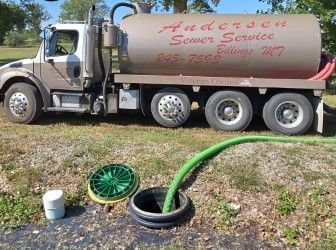 A tan vacuum truck parked on grass with a green hose inserted into an open, round septic access pipe.