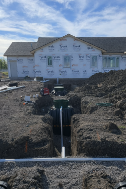 A house under construction with Tyvek house wrap, featuring an excavated yard with a black septic tank and drainage pipes.