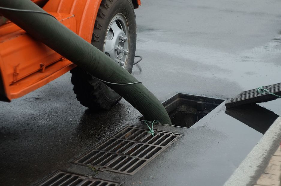 A large hose from an orange utility vehicle is inserted into an open storm drain on a wet asphalt street.