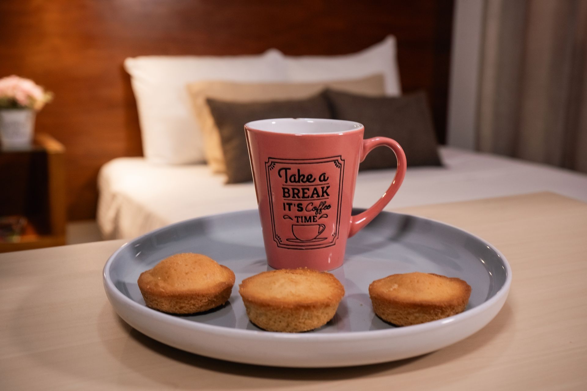 Una taza rosa con el texto “Tómate un descanso”, tres muffins en un plato, colocados sobre una mesa frente a una cama.