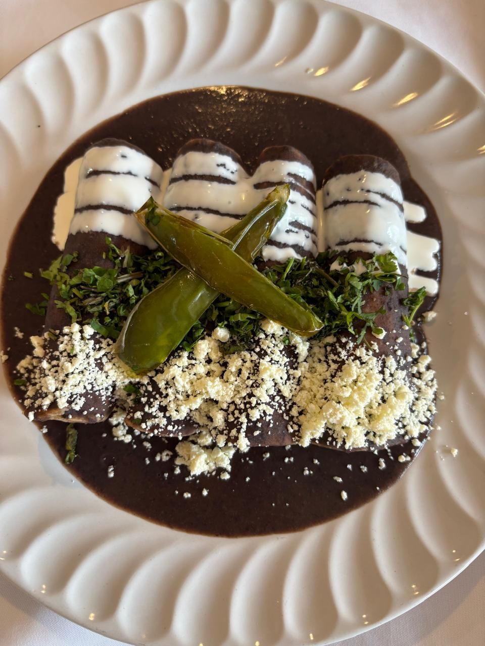 Enchiladas con salsa de mole oscuro, crema blanca, queso, hierbas y pimientos en un plato blanco.