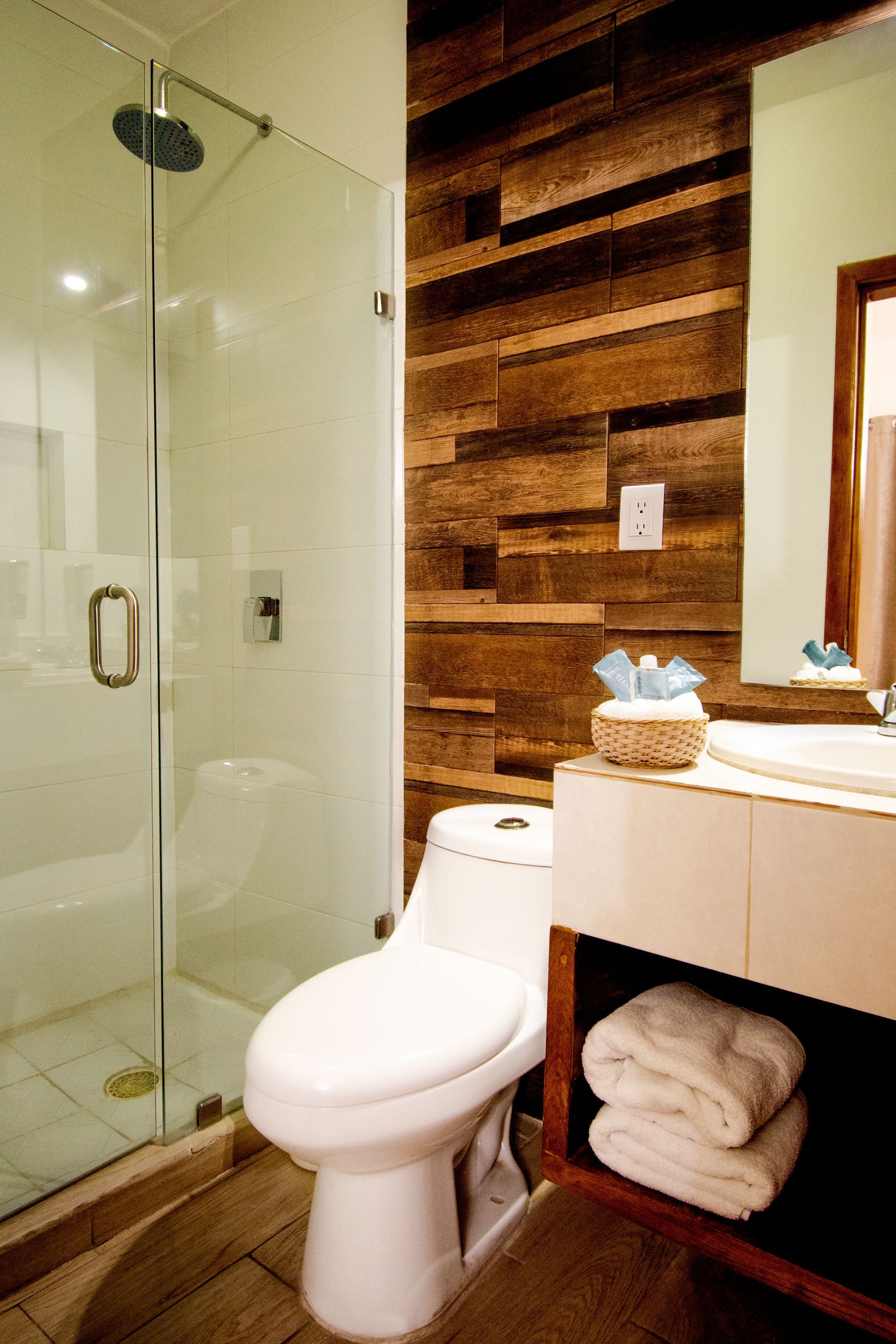 Baño con ducha de cristal, pared revestida de madera, inodoro blanco y tocador con toallas.