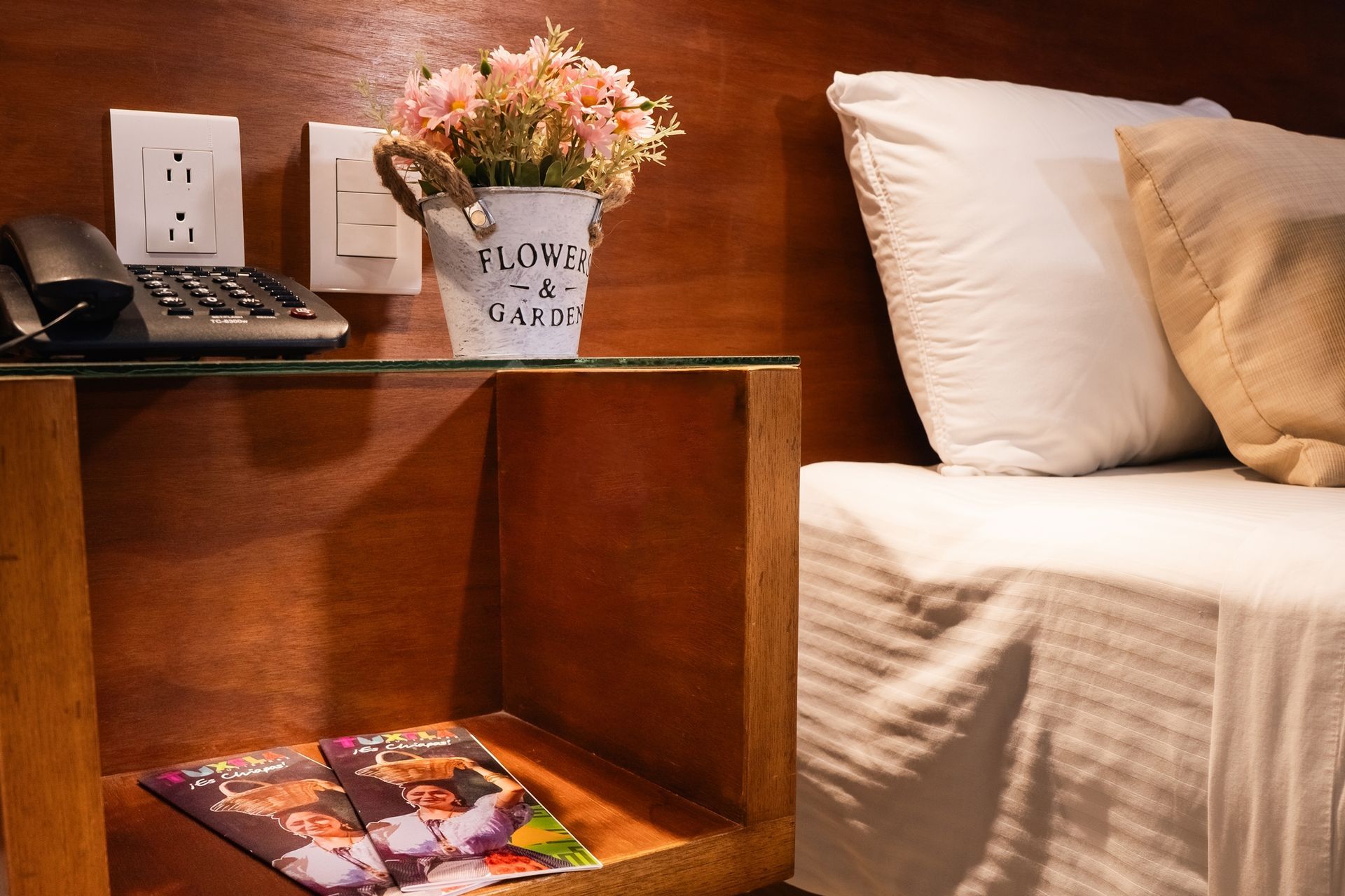 Mesilla de noche con flores, teléfono y folletos junto a una cama con almohadas.