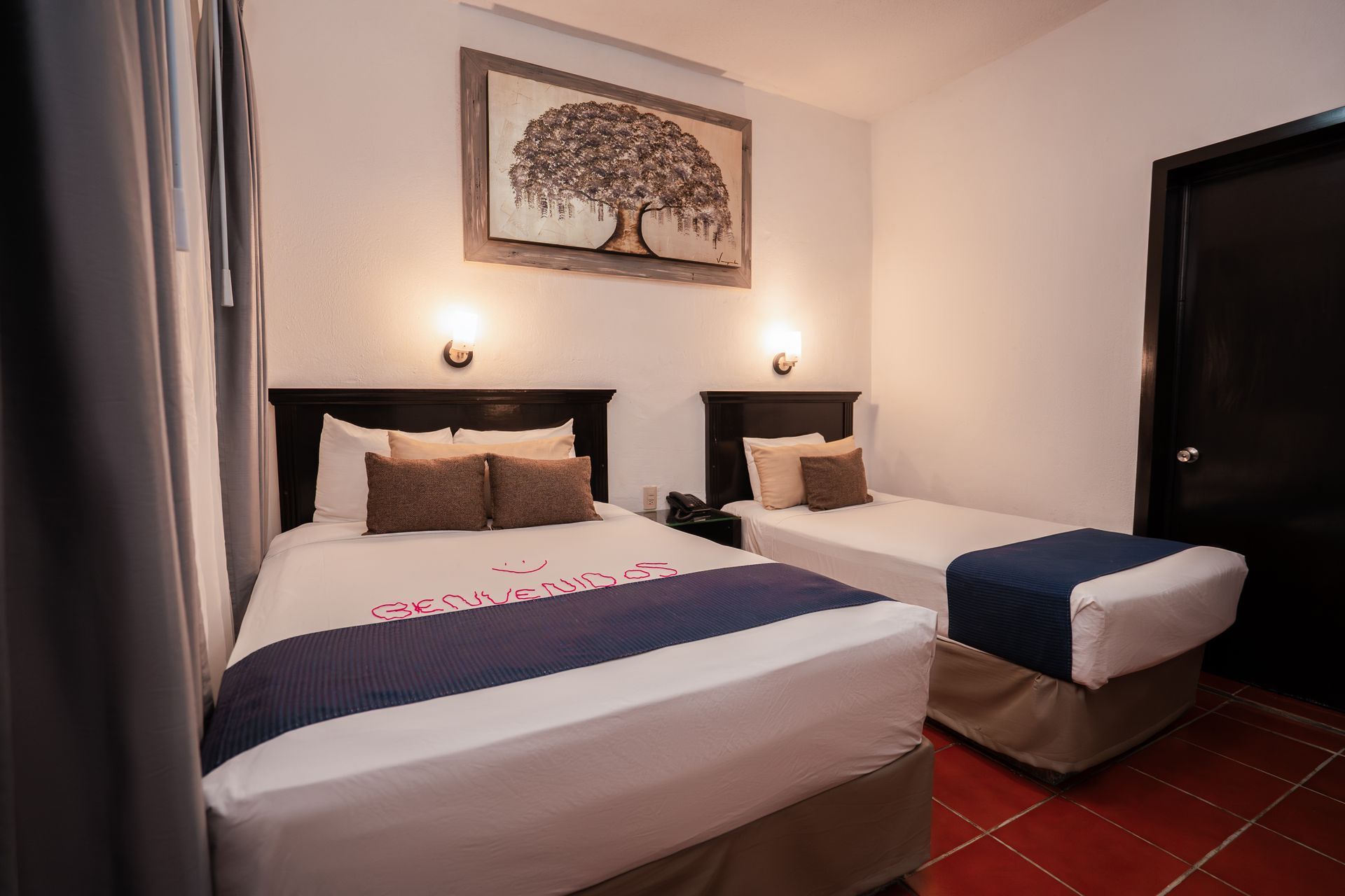 Una habitación de hotel con dos camas, un cuadro de árbol enmarcado y dos apliques de pared.