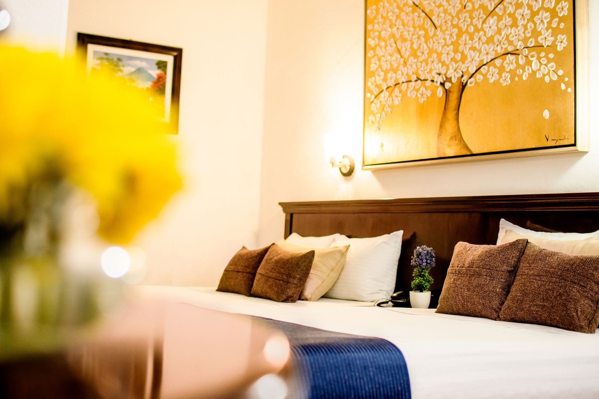 Dormitorio acogedor: cama con almohadas, cuadro dorado, jarrón con flores amarillas, cabecero oscuro.