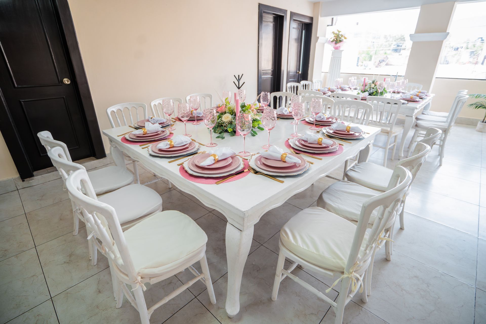 Mesa blanca puesta para fiesta, con platos rosas y dorados, rodeada de sillas blancas, en una habitación luminosa.