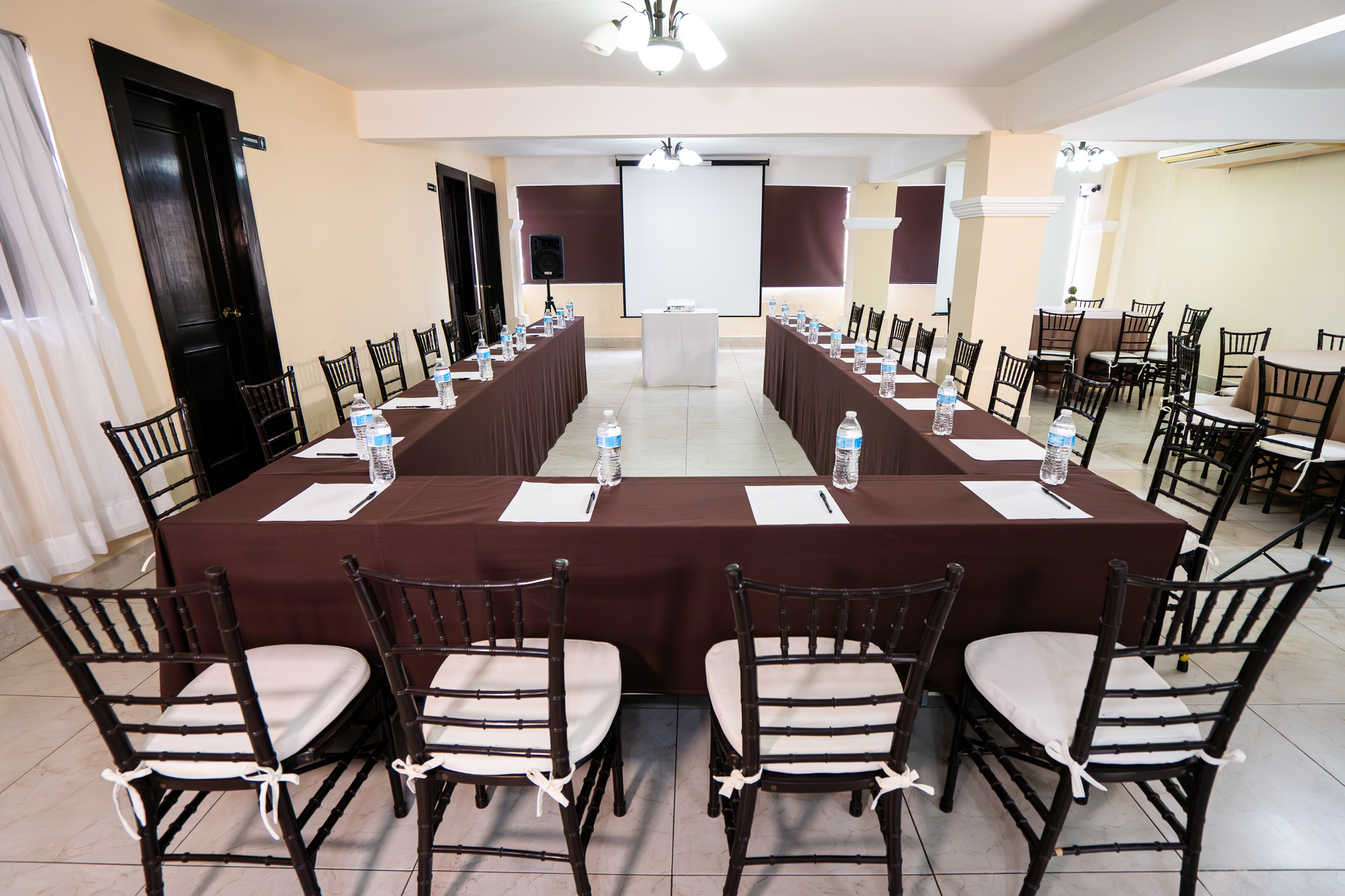 Sala de conferencias con mesa en forma de U, sillas, pantalla de proyección y botellas de agua.