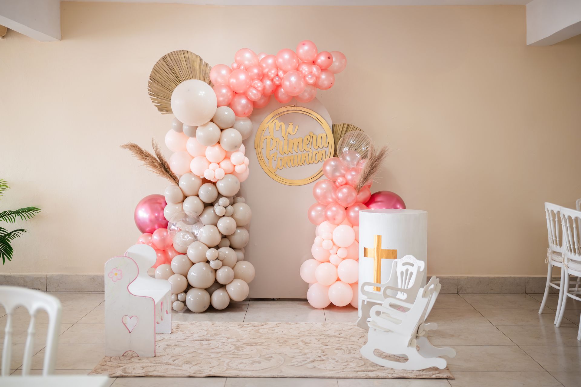Arco decorado con globos rosas y neutros, una cruz blanca y una mecedora en una celebración de bautizo.