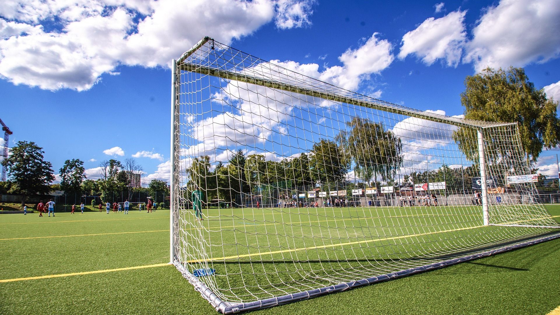 A close-up of a soccer goal net on a green field under a bright, partly cloudy blue sky.