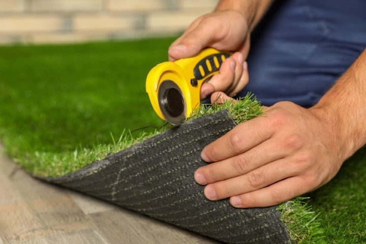A person uses a yellow rotary cutter to trim the edge of a piece of artificial turf resting on a tiled floor.
