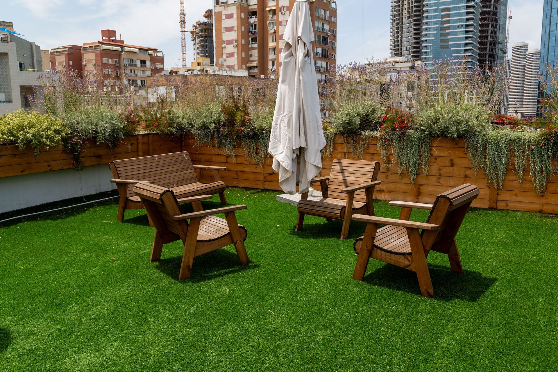 A rooftop terrace with a green artificial turf floor, wooden furniture, a closed umbrella, and planters against a fence.