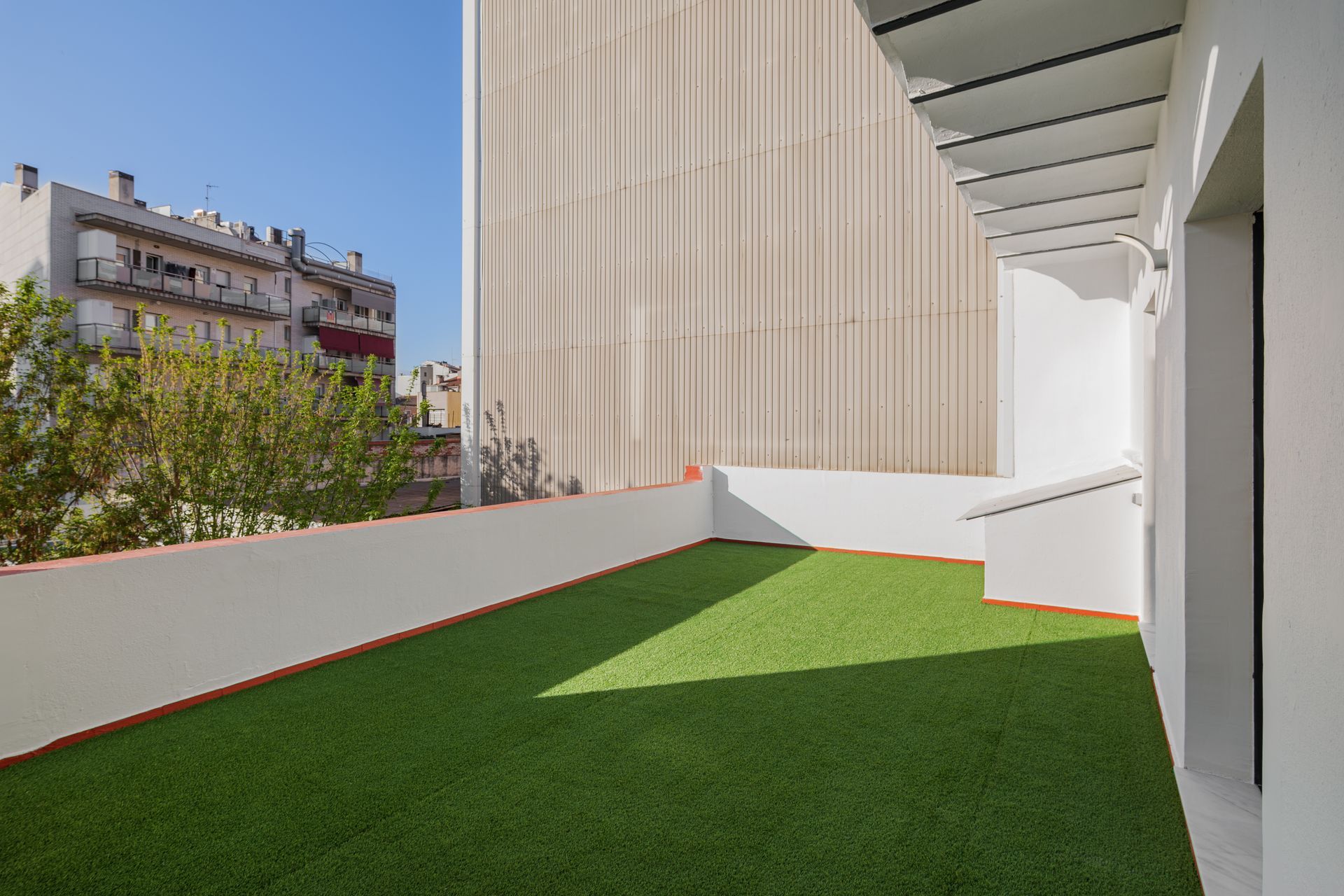 A rooftop balcony with artificial turf, white walls, and an overhead trellis, overlooking a city building and tree.