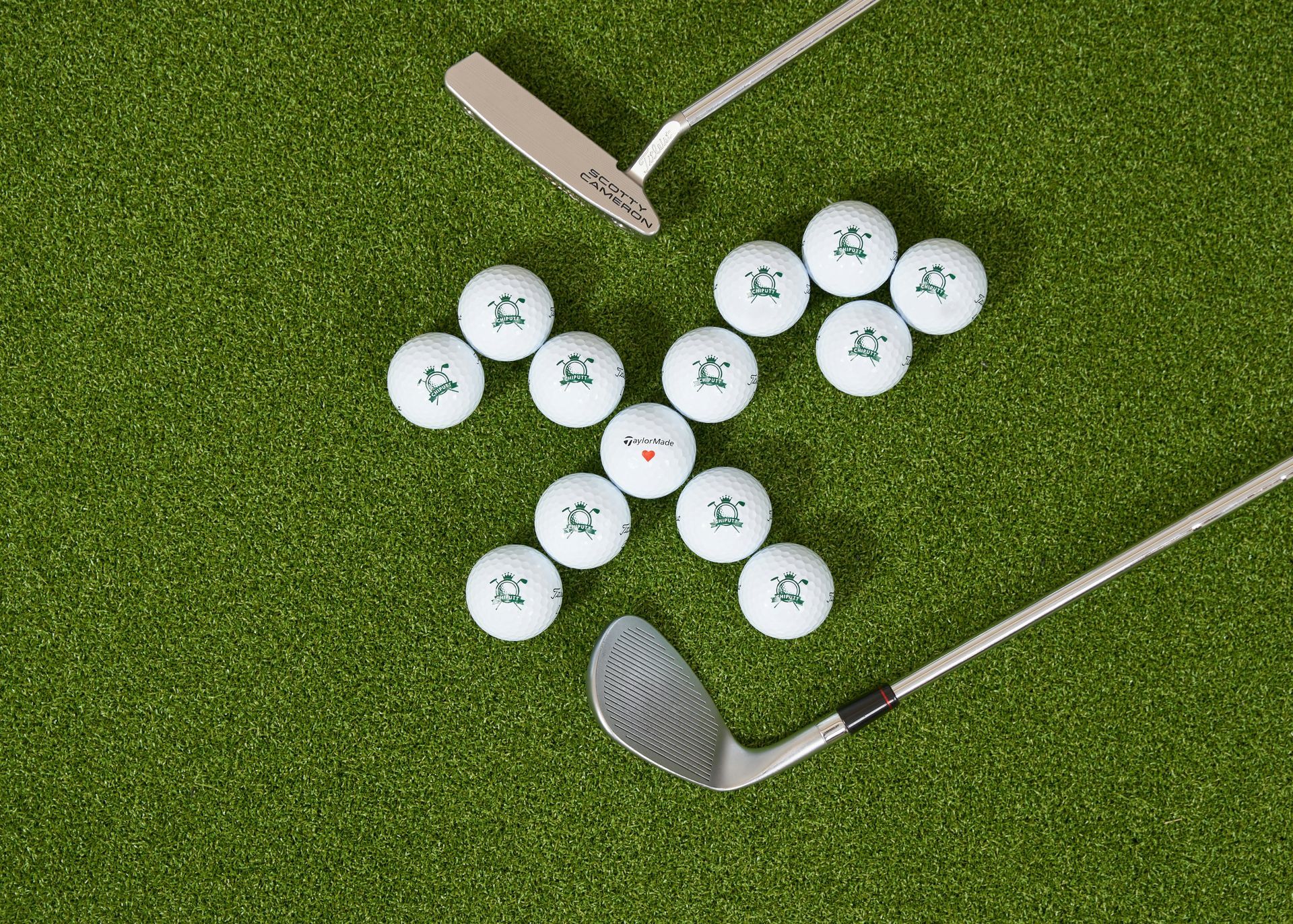 Thirteen white golf balls arranged in a cross shape on green turf with two golf clubs placed nearby.
