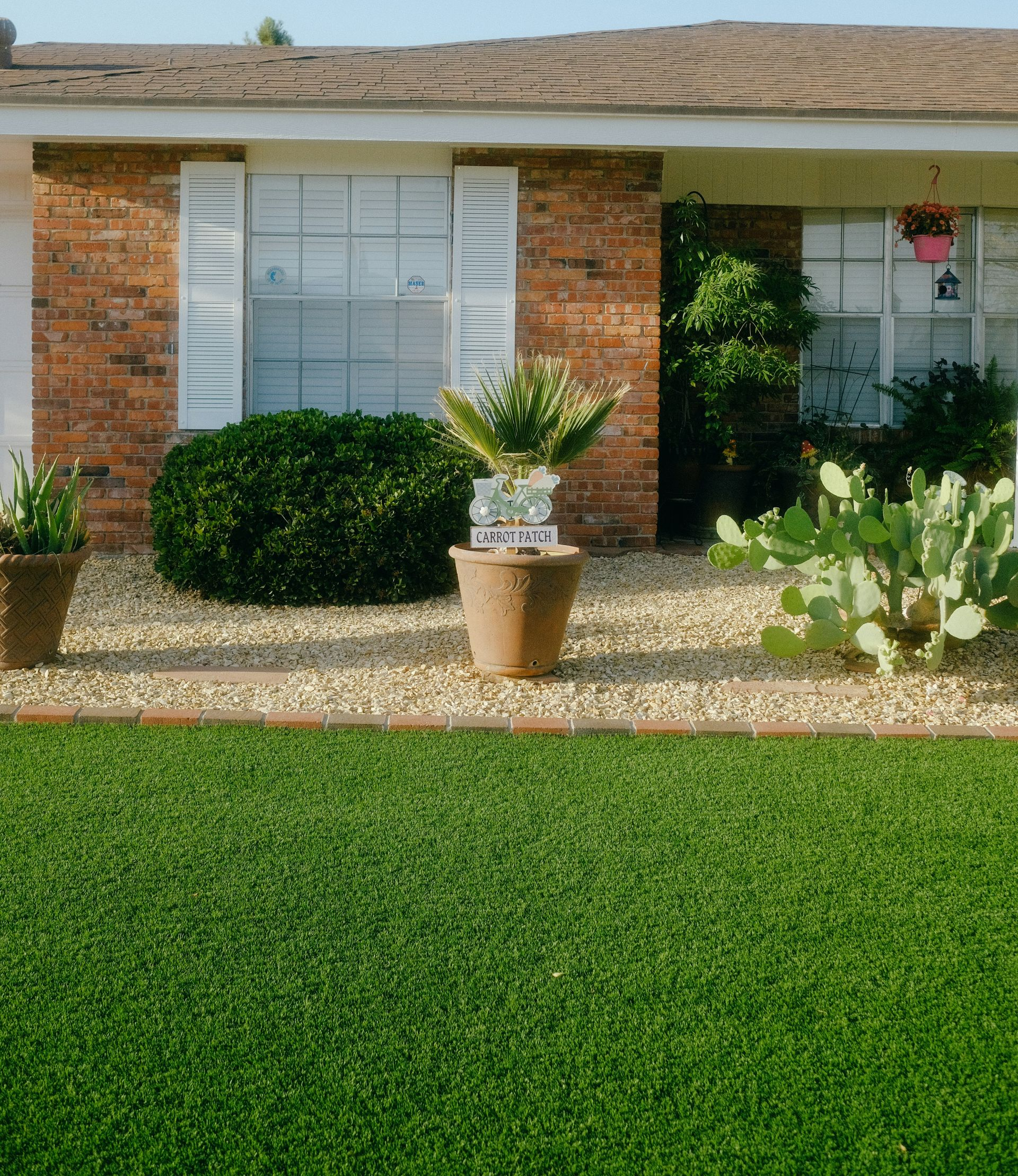 A brick house exterior features a lush green lawn, a rock garden, potted plants, and a small prickly pear cactus.