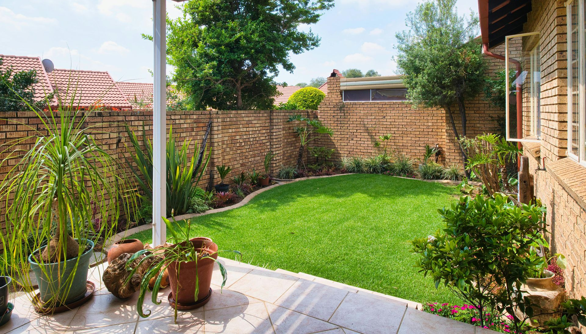 A small, green lawn is bordered by brick walls, potted plants, and a tiled patio under a covered porch.