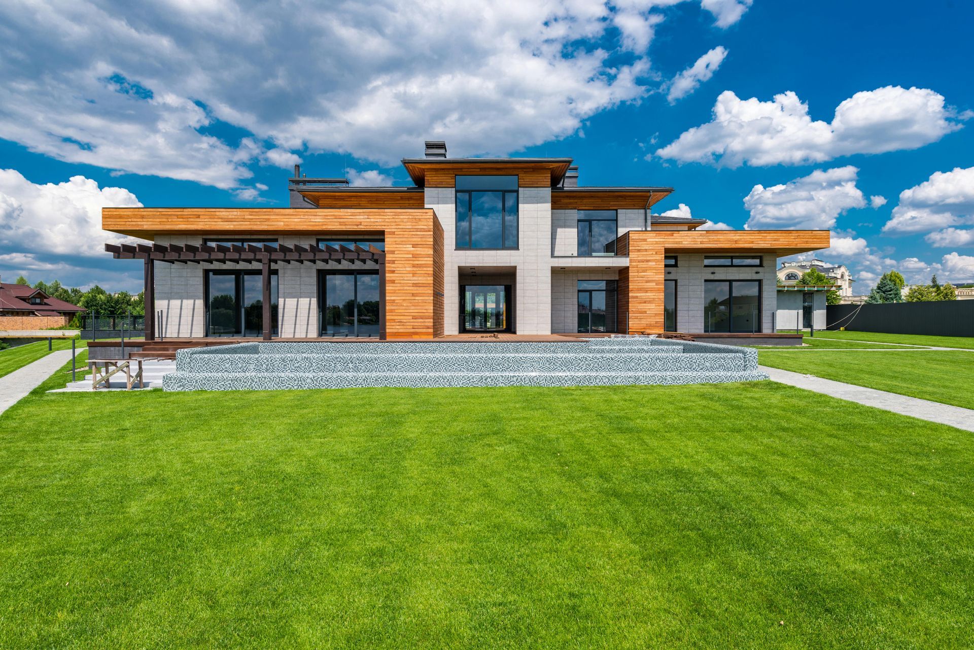 A modern two-story home with wood paneling and stone textures, featuring large glass windows, overlooking a green lawn.