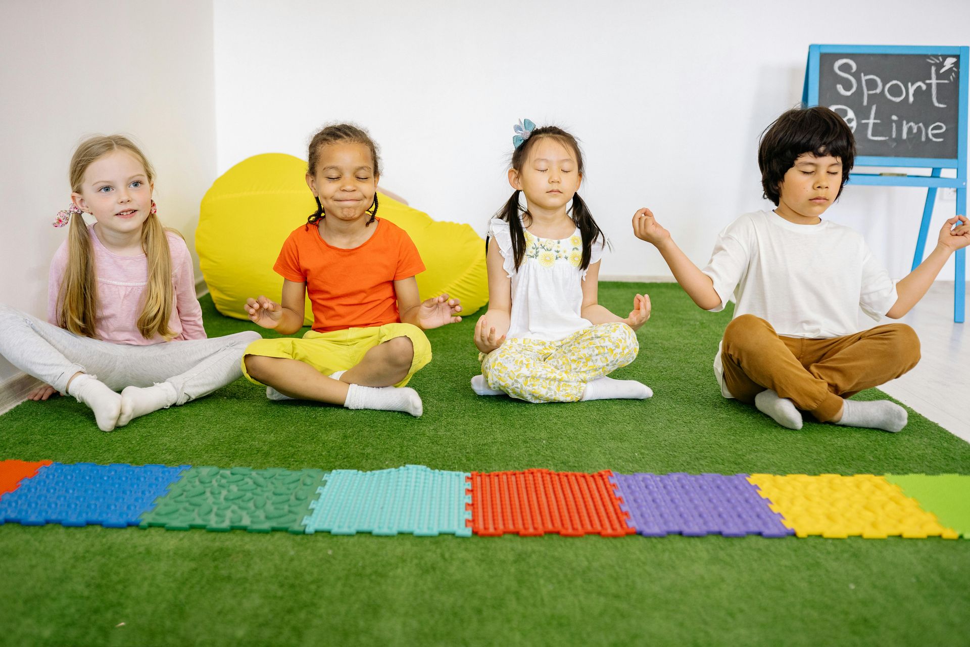 Four children sit in meditation poses on an artificial grass floor in a classroom with a colorful mat and a sport sign.