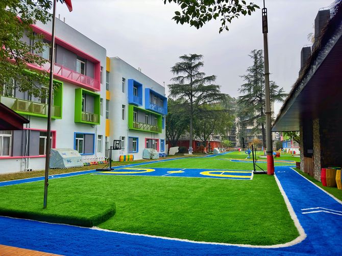 A brightly colored modern building overlooks a school playground with artificial green turf and a blue running track.
