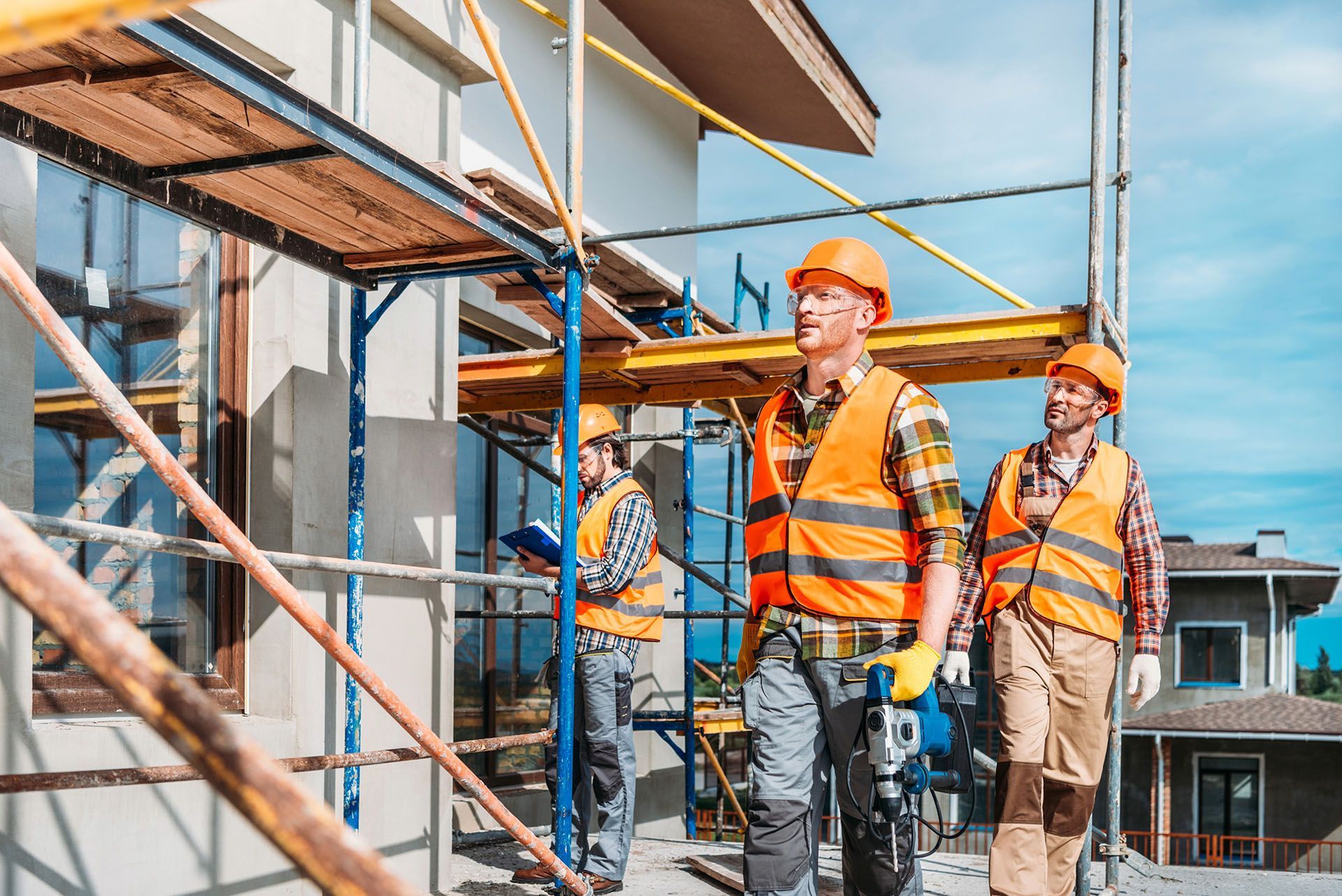 Construction workers in orange vests and hard hats walk past scaffolding on a building.