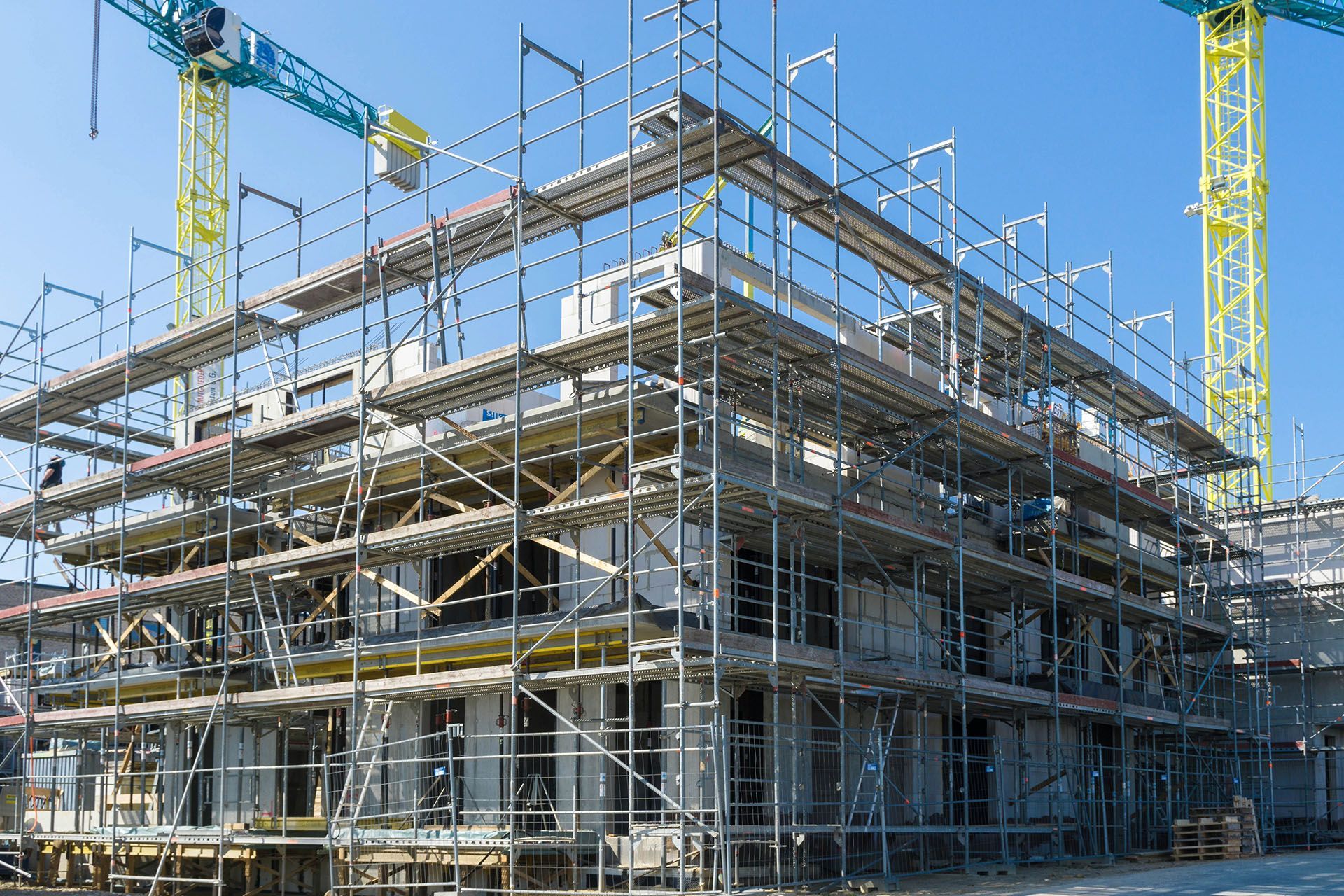 Construction site with scaffolding surrounding a multi-story building, cranes in background, clear blue sky.