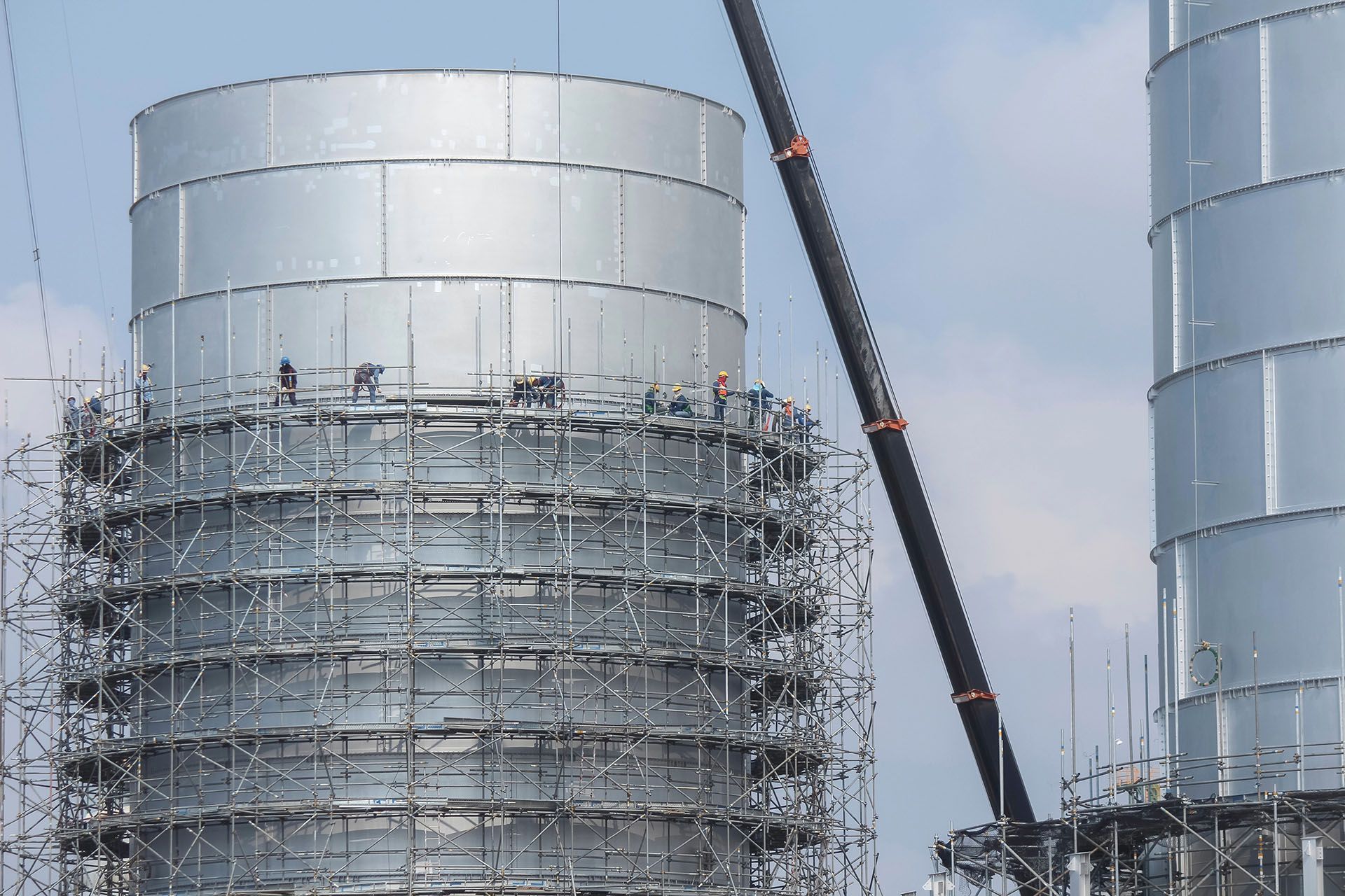 Construction workers on scaffolding build a large, silver industrial tower.