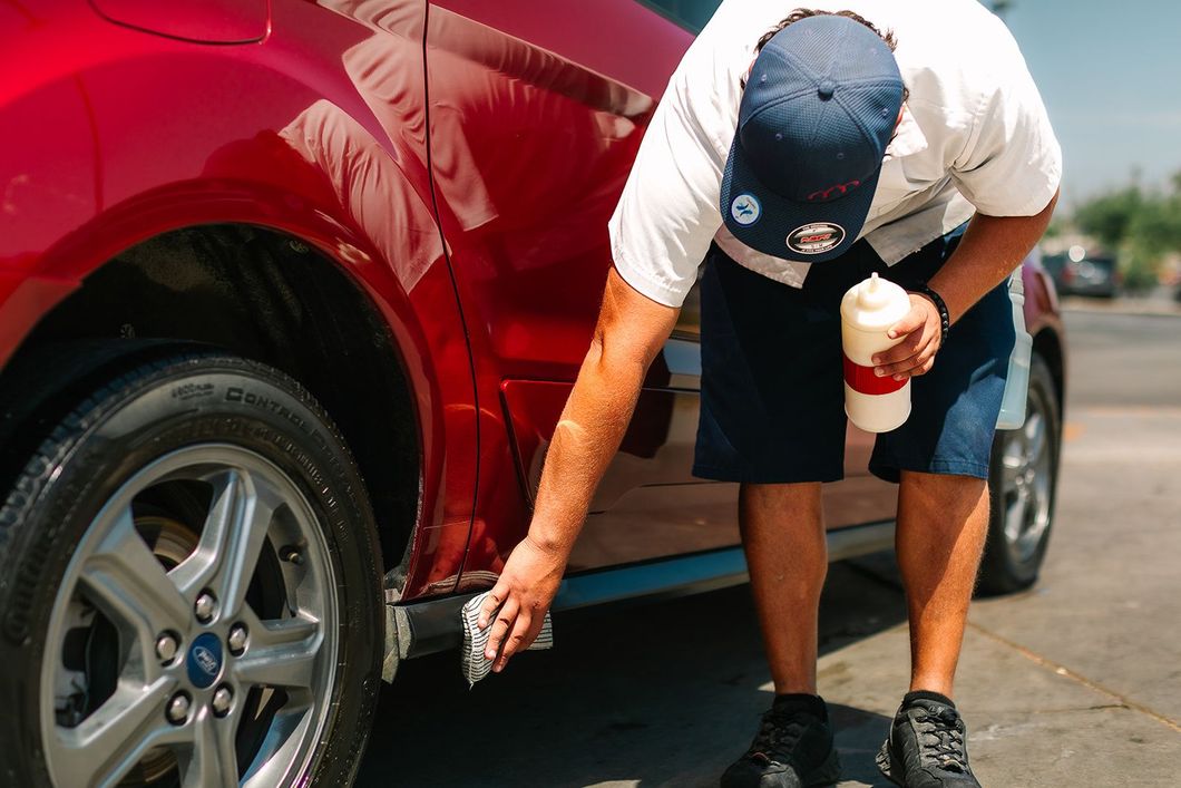 Person washing a red car with a sponge and a bottle of soap outdoors.