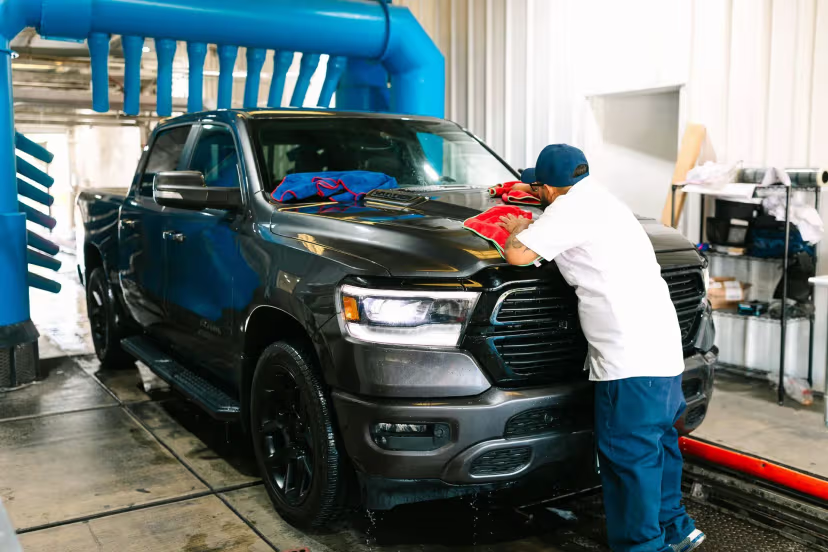 A person wiping a dark truck at a car wash. The wash equipment is visible above.