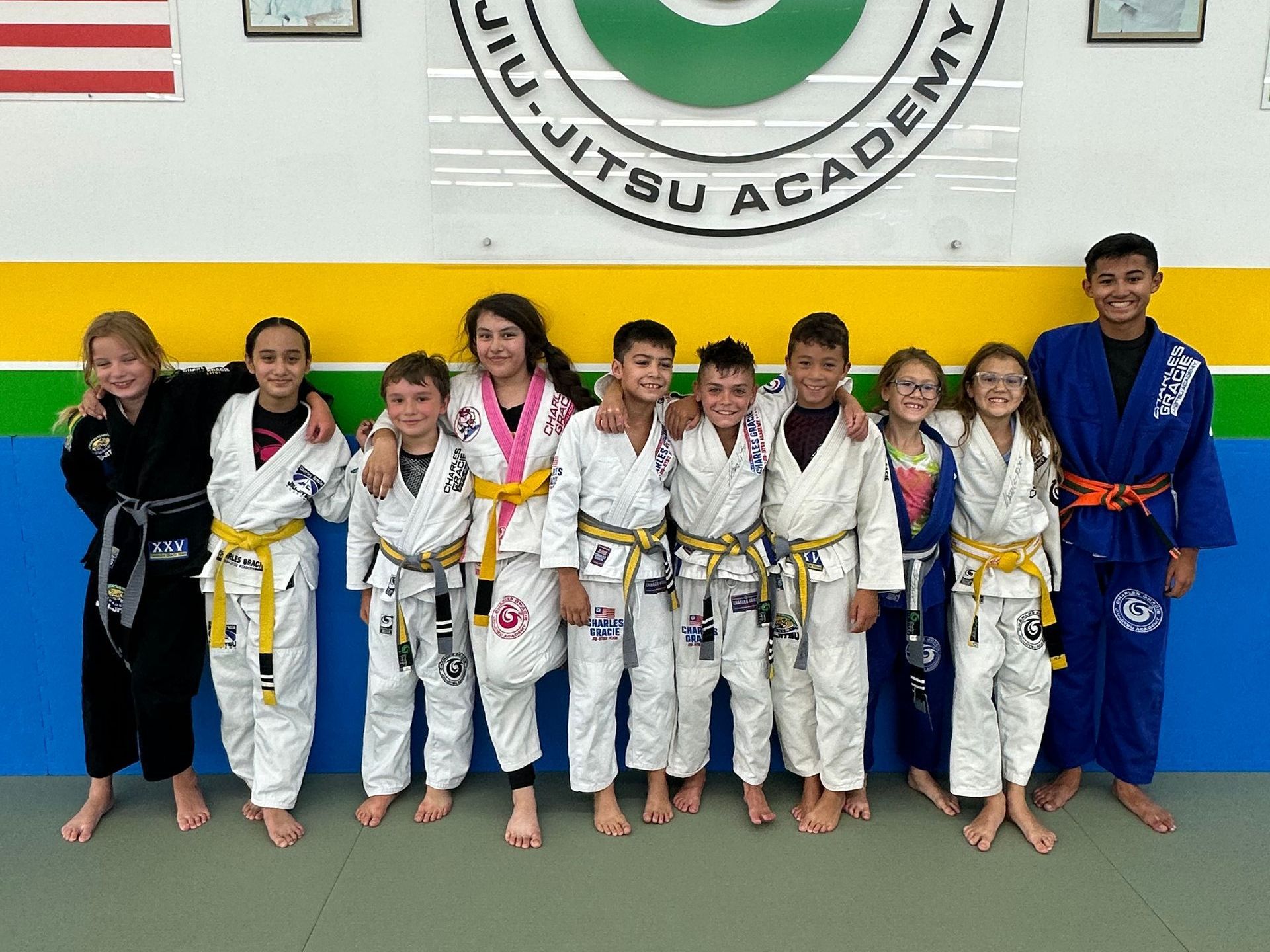 Group of children in Jiu-Jitsu uniforms, smiling, posing together in front of academy logo.