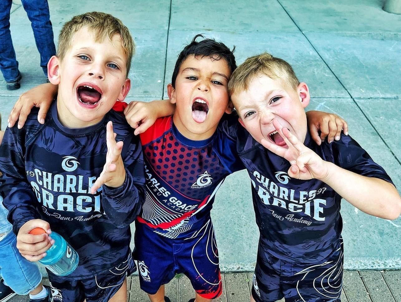 Three young boys in athletic gear cheer, arms around each other, outdoors.