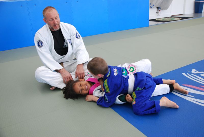 Man in gi watches two children in gis play on mat; one on top, one on bottom, all smiling.