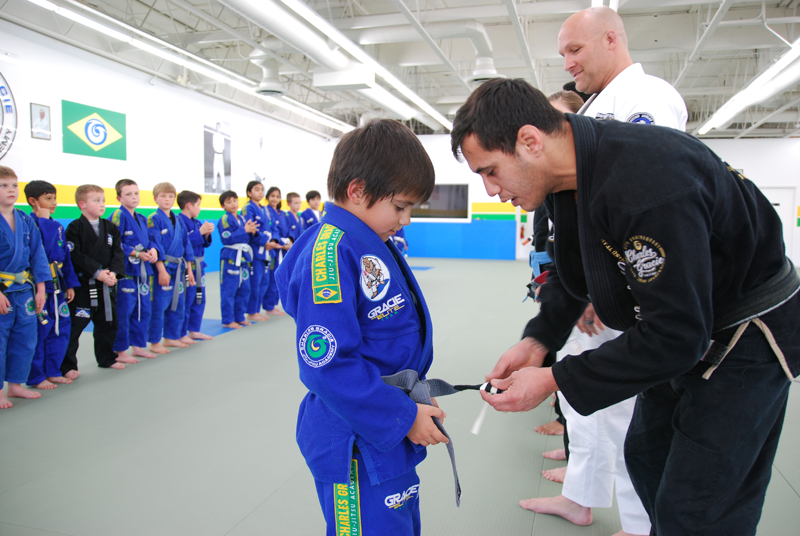 Child in blue gi getting belt tied by instructor; other students in the background.