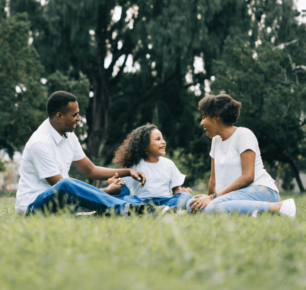 A family sits on the grass in a park, laughing and interacting with one another while wearing matching white shirts.