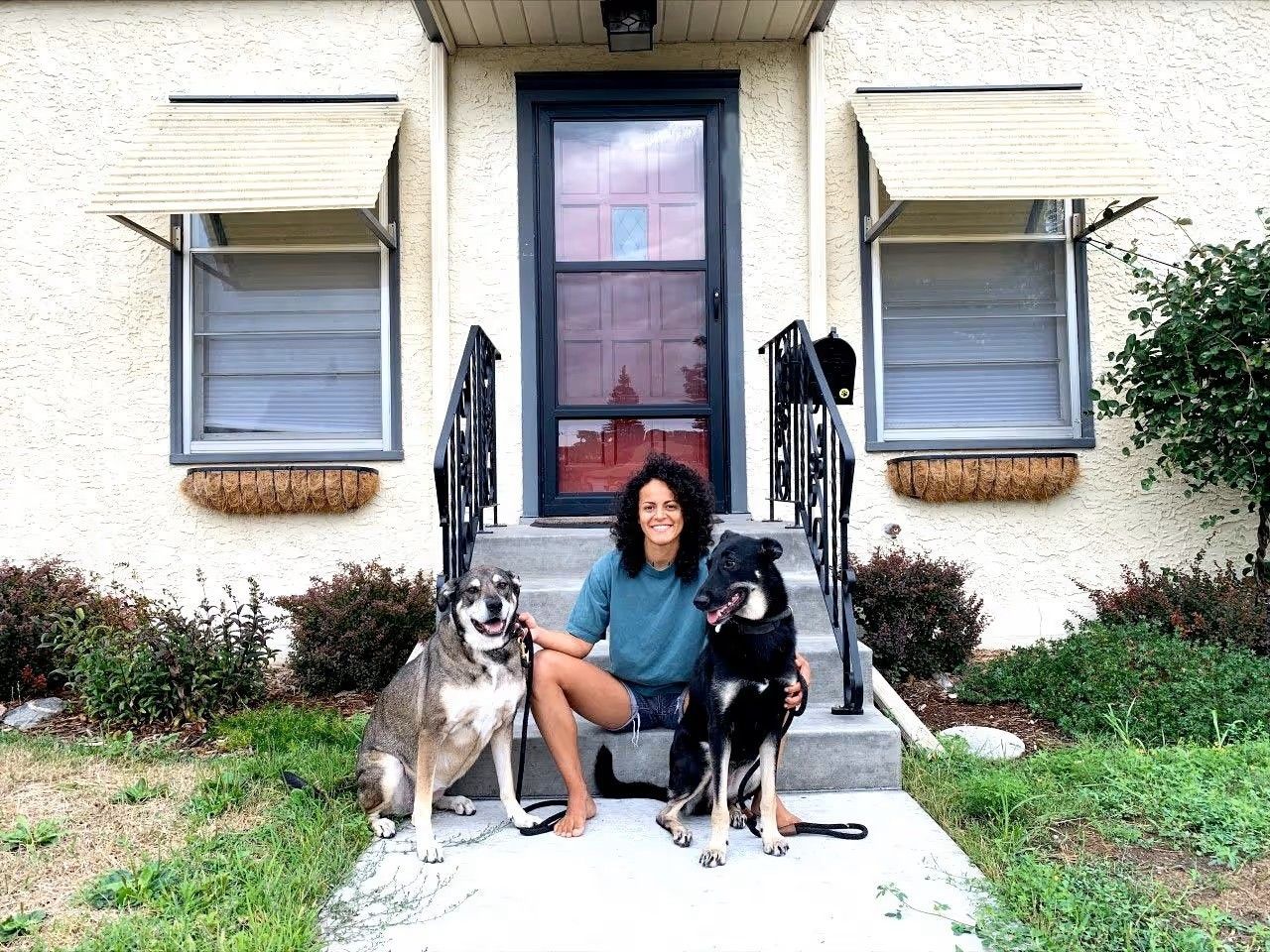 A person sits on concrete front steps of a house with two large dogs on leashes, one on each side.